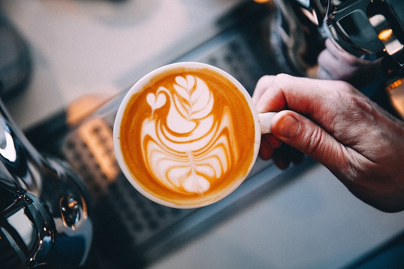 Barista's hand holding cappuccino with intricate latte art design showing swan or tulip pattern in foam